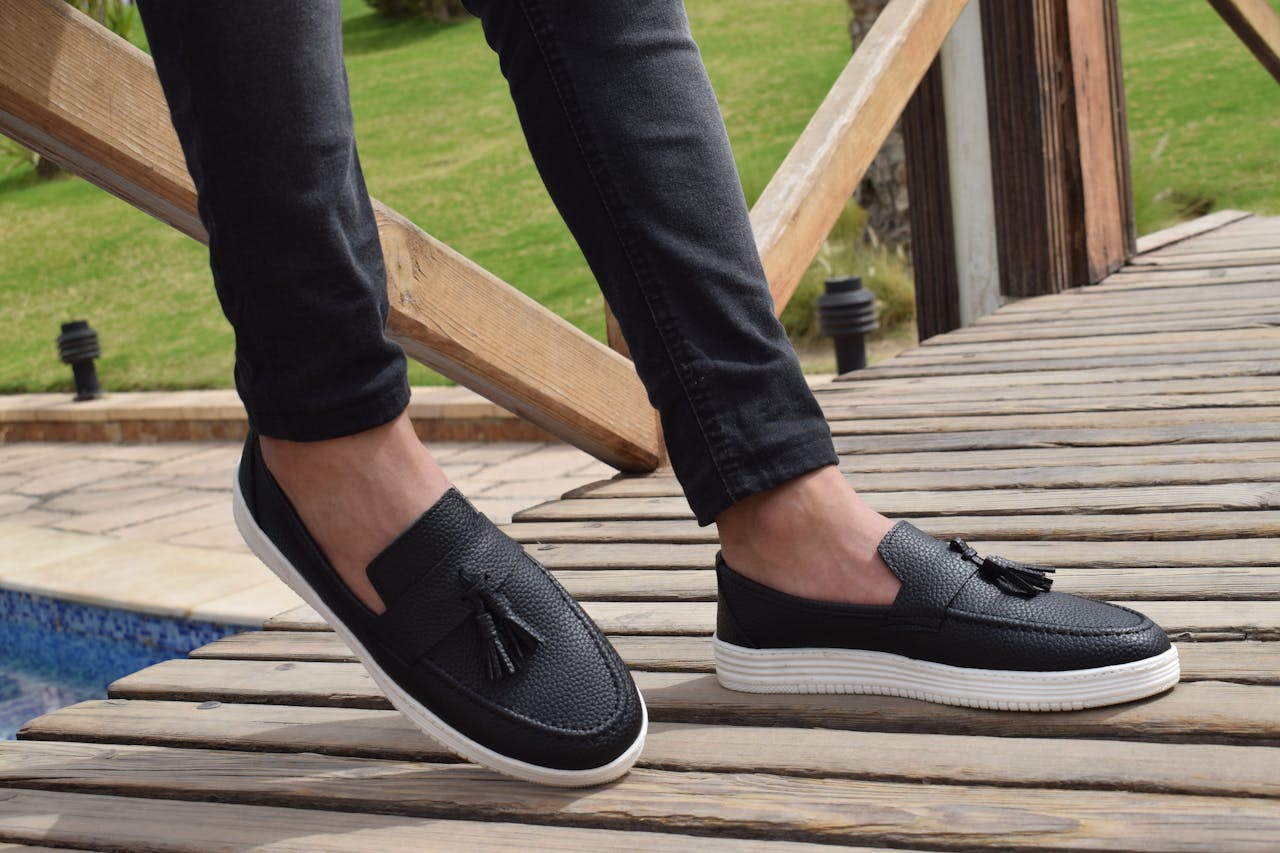Stylish black tassel loafers worn by a person standing on a wooden deck. Perfect for outdoor leisure fashion.